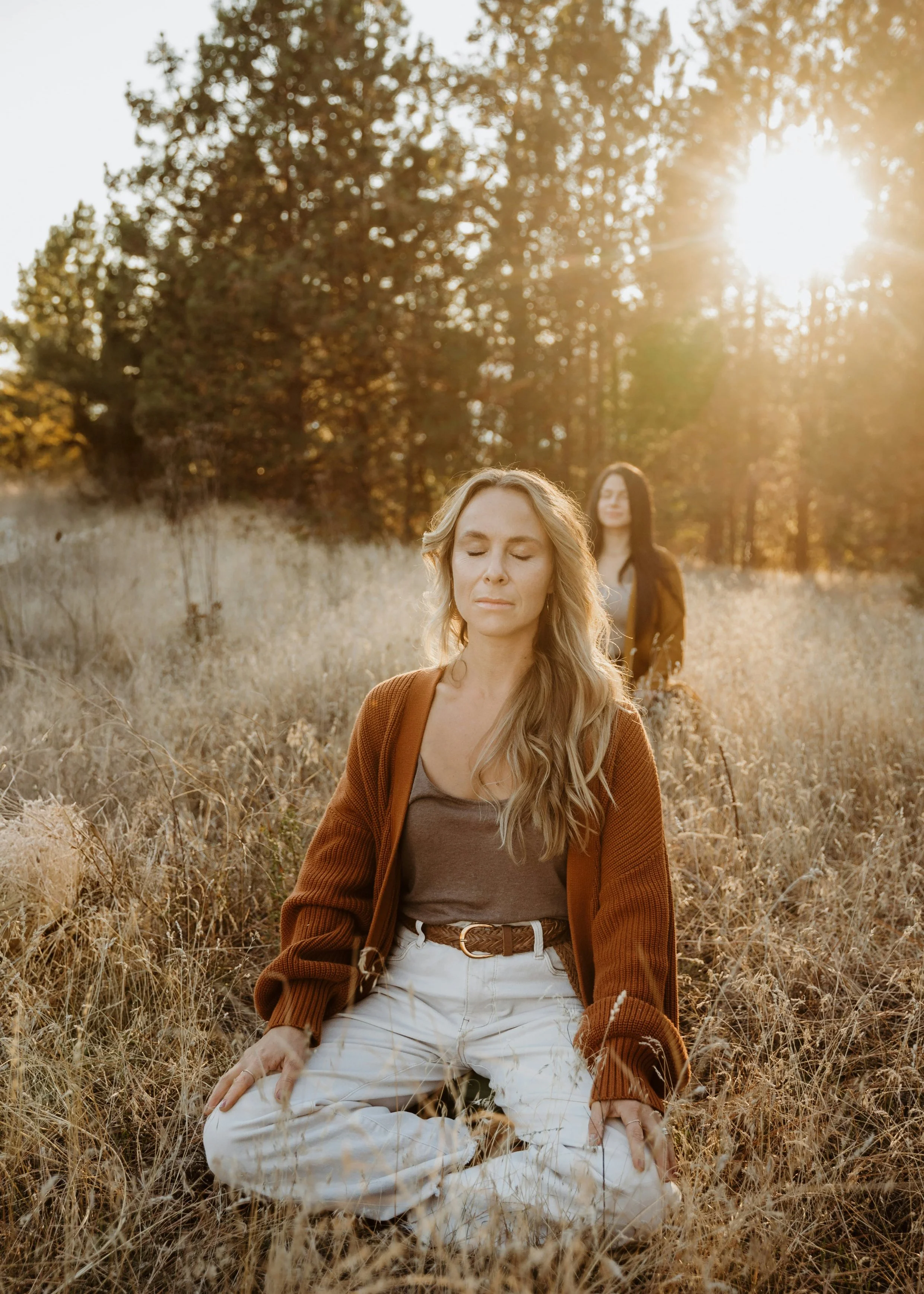 Two women meditating outdoors in a grassy field during sunset, with trees in the background.