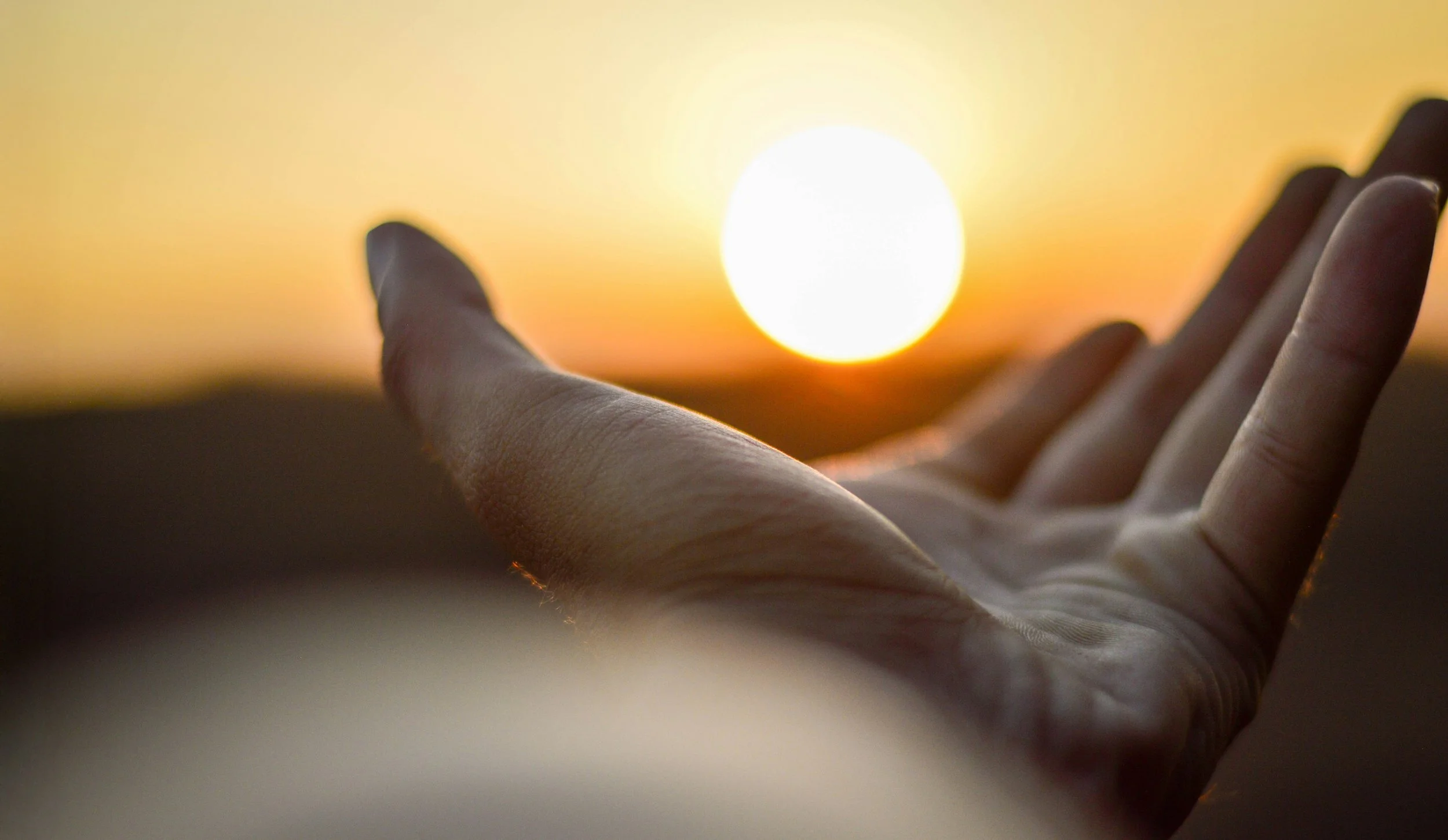 Close-up of a hand reaching toward the setting or rising sun on the horizon, creating a silhouette against the vibrant sky with warm orange and yellow hues.