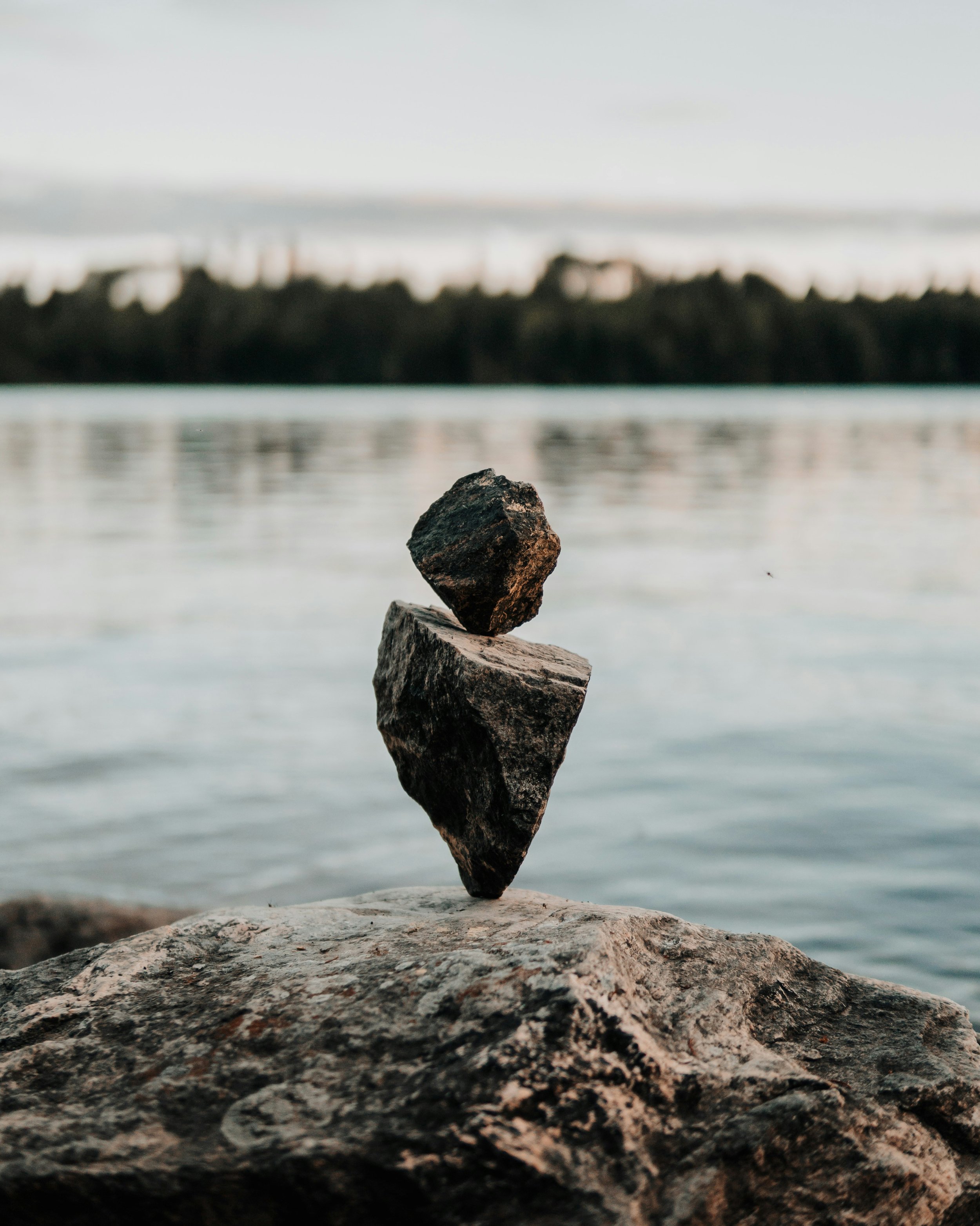 Three rocks stacked on top of each other on a rocky shoreline with water and blurred trees in the background.