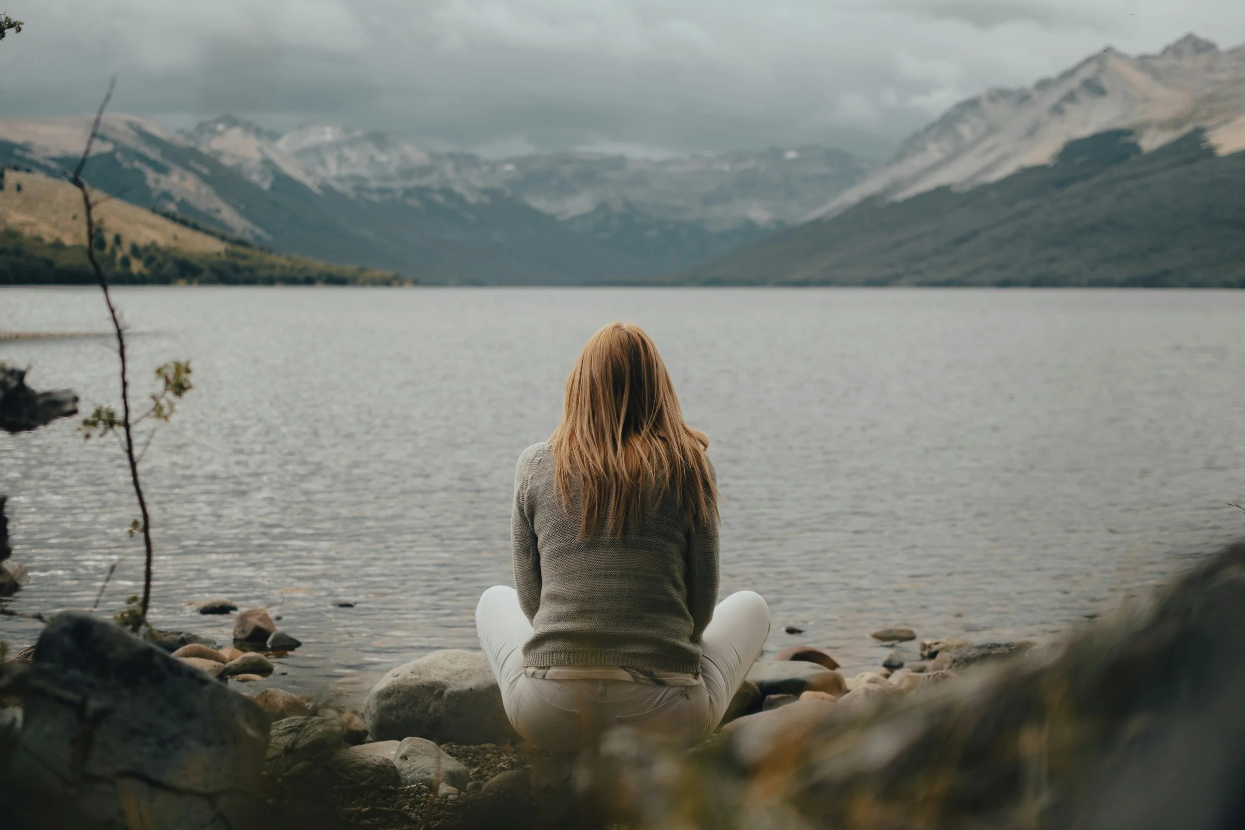 A woman sitting on rocks by a lake, facing mountains under a cloudy sky.