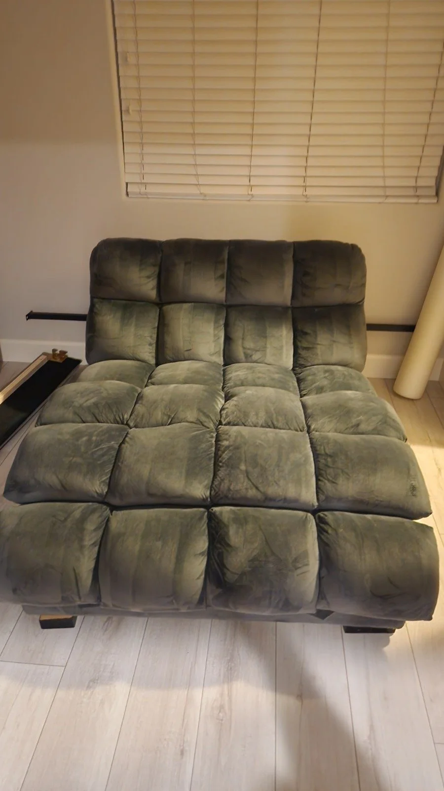 A green velvet sectional sofa with a tufted design placed in front of a window with closed blinds, on a light wooden floor.