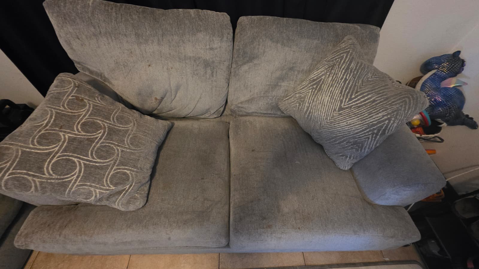 A beige fabric sofa with three cushions. The cushions are patterned, with one having a circular design, and another with a zigzag pattern. The sofa appears worn and stained, with a dark curtain behind it and various items to the right side.
