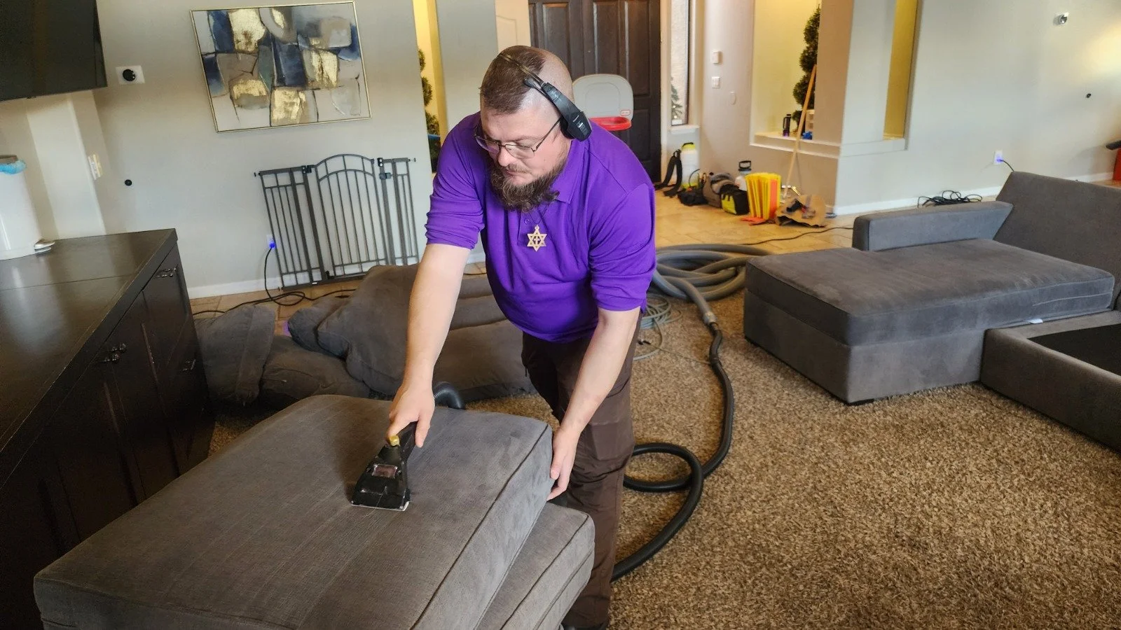 A man steam cleaning a gray upholstered ottoman in a living room, wearing a purple shirt, glasses, headphones, and a star-shaped necklace, with furniture and cleaning supplies around him.