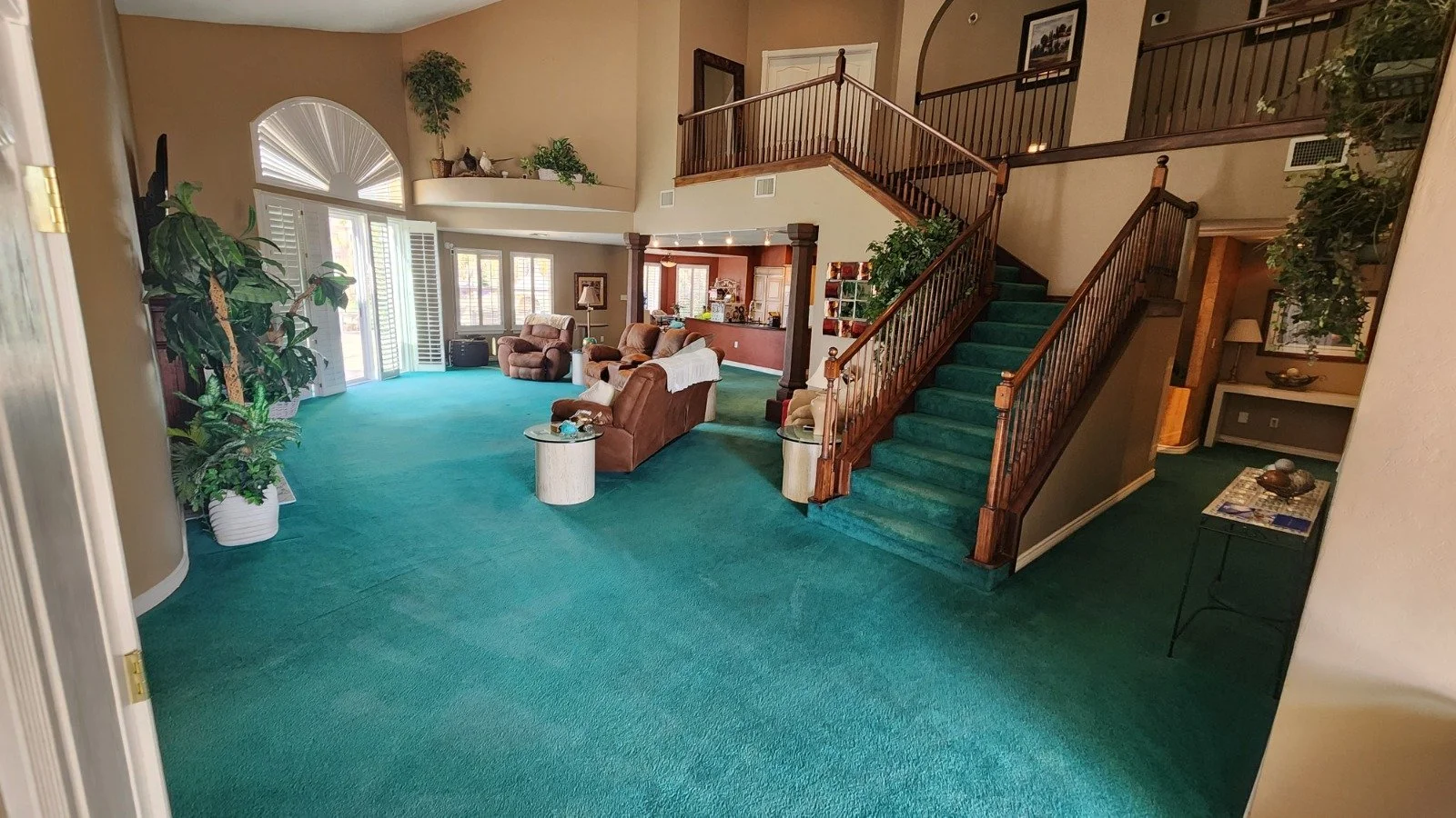 Interior of a spacious living room with green carpet, multiple sofas, large windows with white shutters, and a wooden staircase with green carpeting leading to the second floor. All carpet was cleaned.