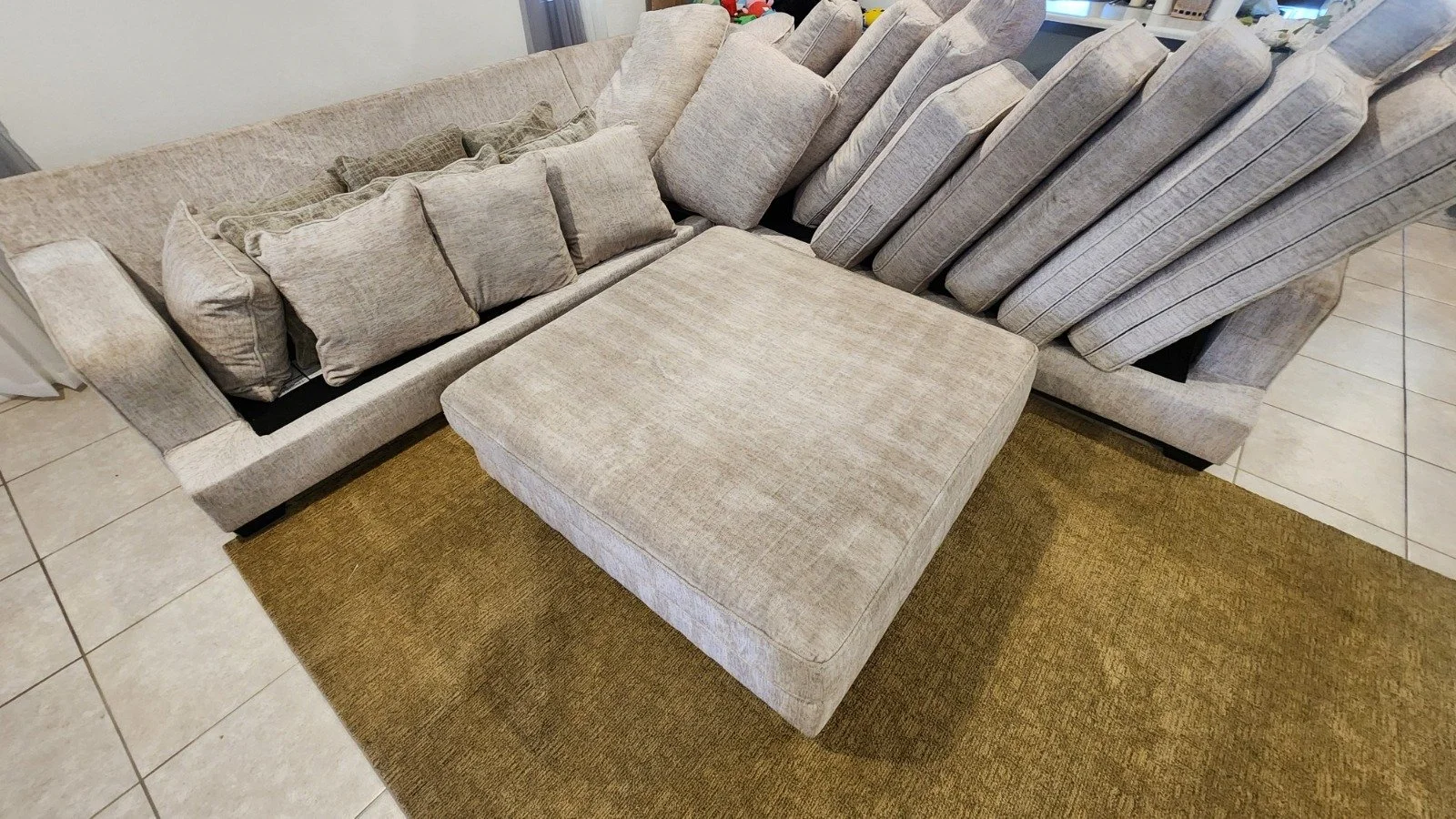 A large beige fabric sectional sofa with multiple cushions, positioned partly on a tan rug and partly on a tiled floor.