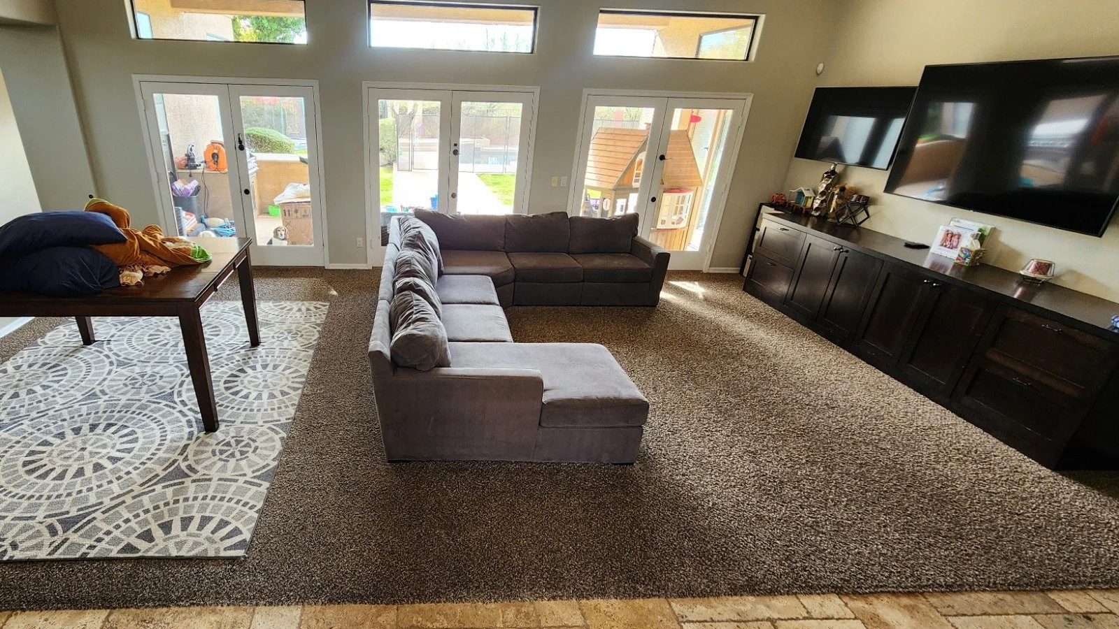 A spacious living room with large windows and glass doors overlooking a backyard. Contains a sectional sofa and carpet that was steam deep cleaned.