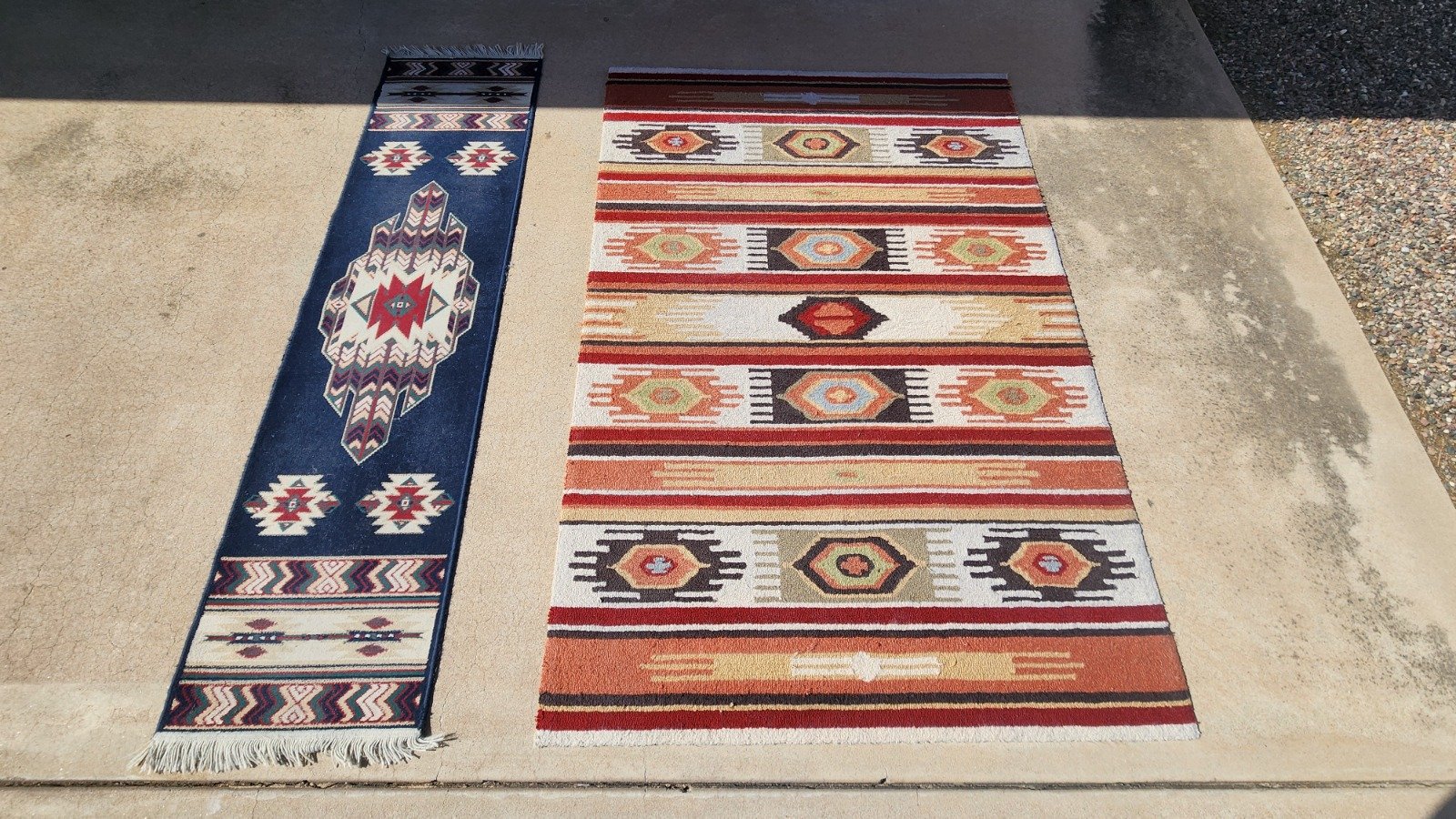 Two colorful woven rugs laid side by side on a concrete patio, one with a navy blue background and geometric patterns, the other with red, beige, black, and green stripes and geometric designs. Both deep cleaned