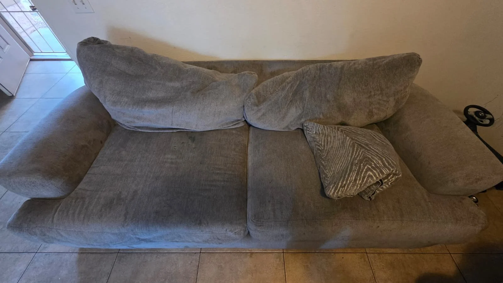 A beige upholstered sofa with three cushions, one of which has a patterned cover, positioned near a wall and a closet door, on a tiled floor.