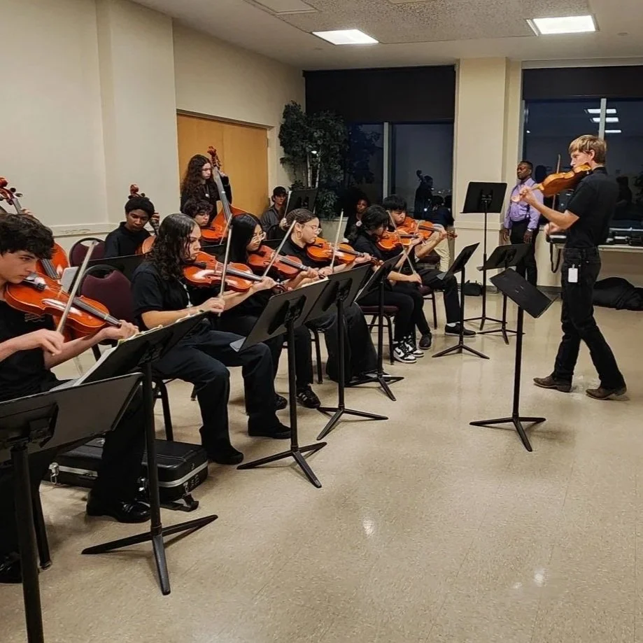 A group of students playing violins in a classroom with a John Shelley standing in front of them.