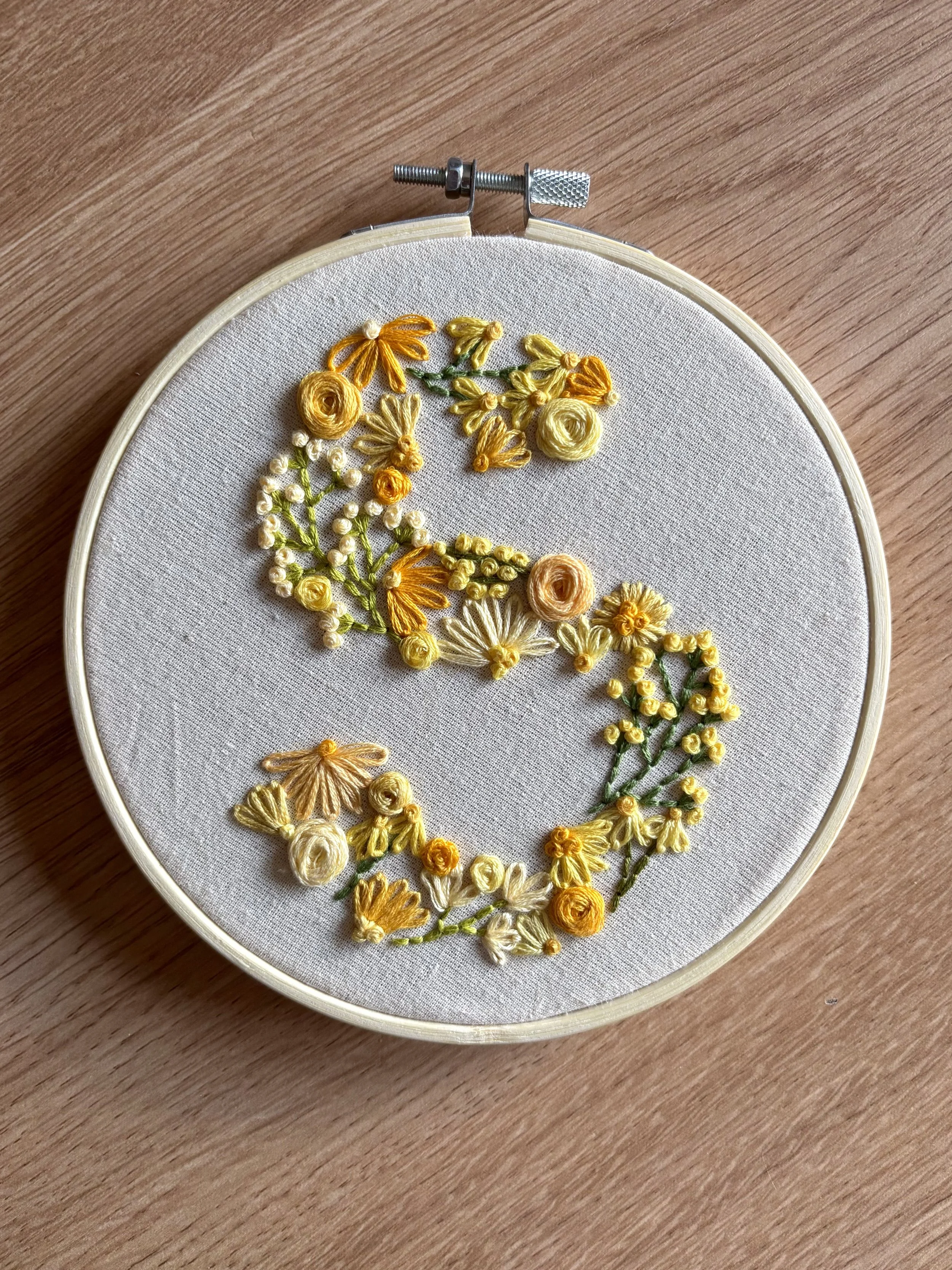 Embroidery hoop with fabric featuring floral embroidery forming the letter S in yellow, gold, white, and green thread.