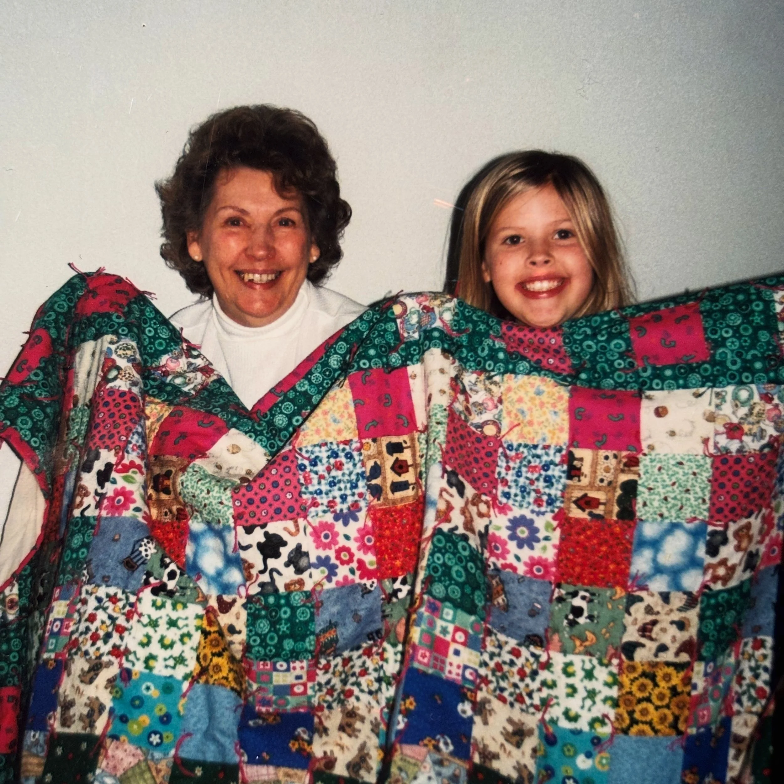 A woman and a young girl smiling behind a colorful quilt made of various patterned fabric squares.