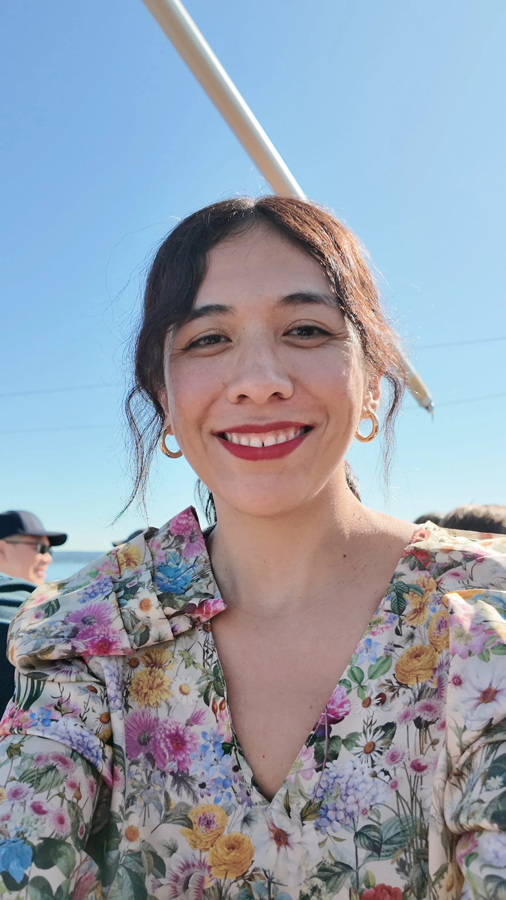 Close-up of a smiling woman with dark hair, wearing gold hoop earrings and a floral shirt, outdoors on sunny day with clear blue sky.