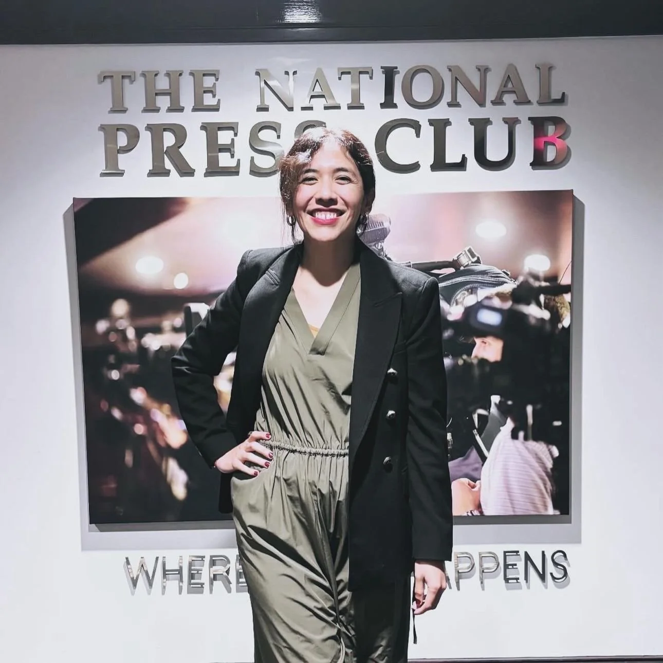 Woman smiling and posing in front of a backdrop at the National Press Club, with camera equipment and flashing lights in the background.