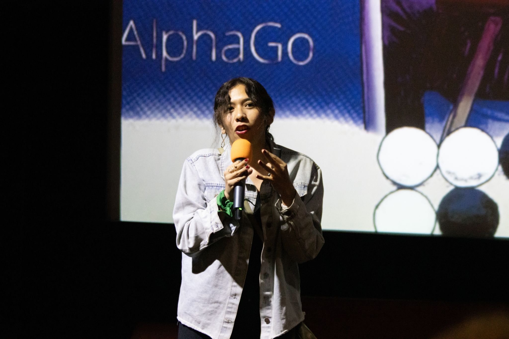 A woman holding a yellow microphone, speaking on stage in front of a projection with the word 'AlphaGo' and images of Go game stones.