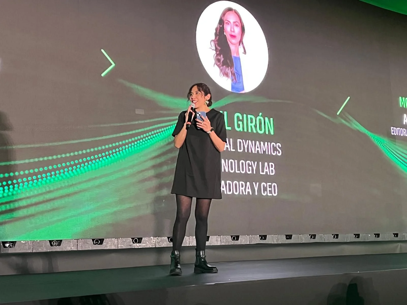 A woman standing on stage holding a microphone, in front of a large digital screen displaying her profile picture, name, and professional titles.