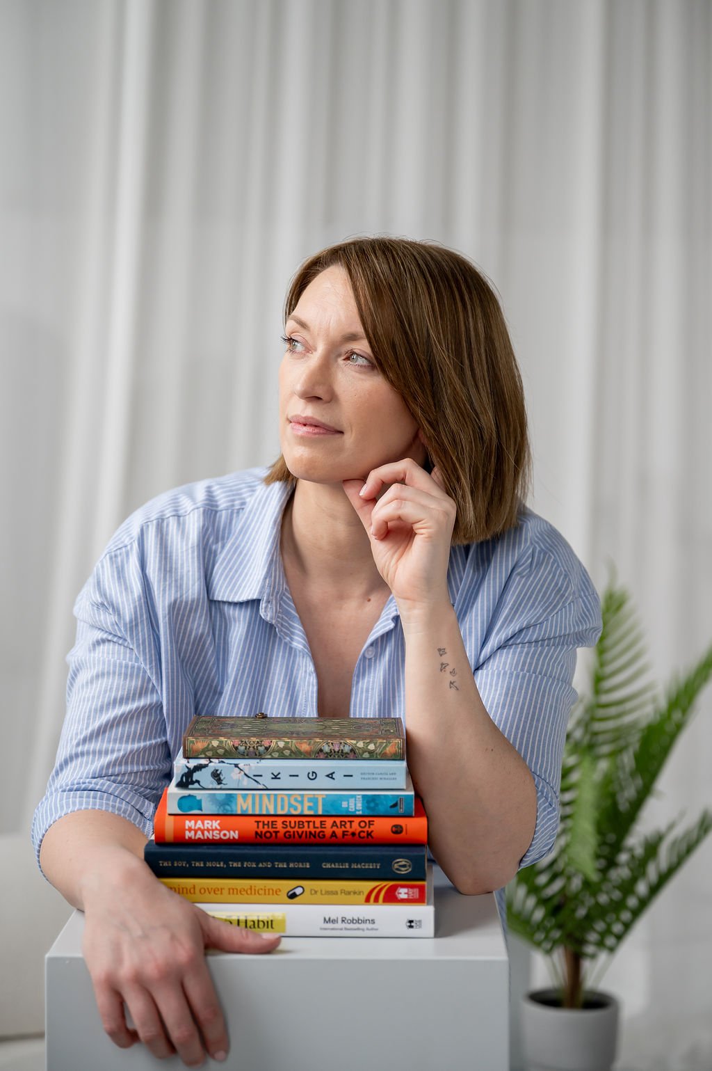 Carla Dawn, coach, wears a blue striped shirt and leans on a stack of books, looking thoughtfully to the side. A green fern is visible in the background.