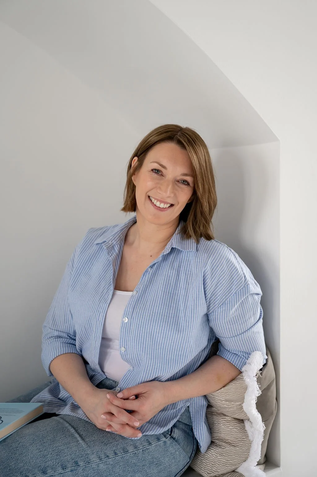 A woman with light brown hair, wearing a blue striped shirt and white top, sits on a cushion against a white wall, smiling at the camera.