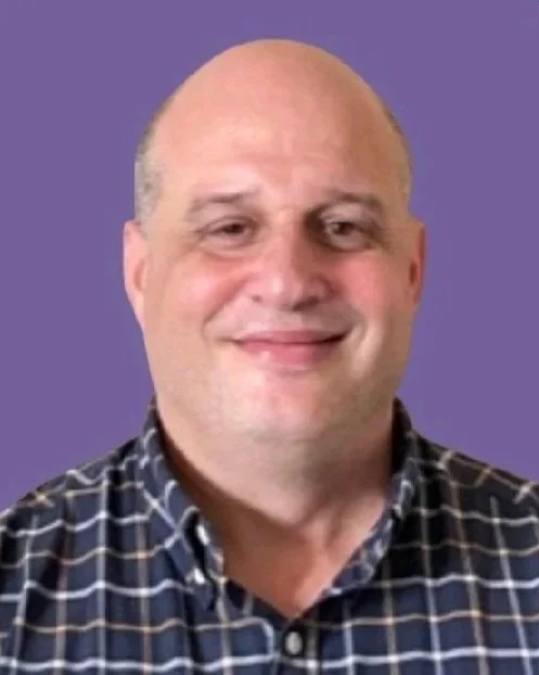 A smiling man with a bald head and wearing a checkered shirt, posed against a purple background.