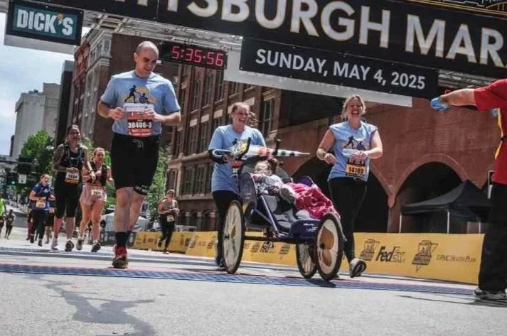People participating in a marathon race, running along a city street with a banner overhead that reads 'Pittsburgh Marathon' and a digital clock showing the time, Sunday, May 4, 2025.