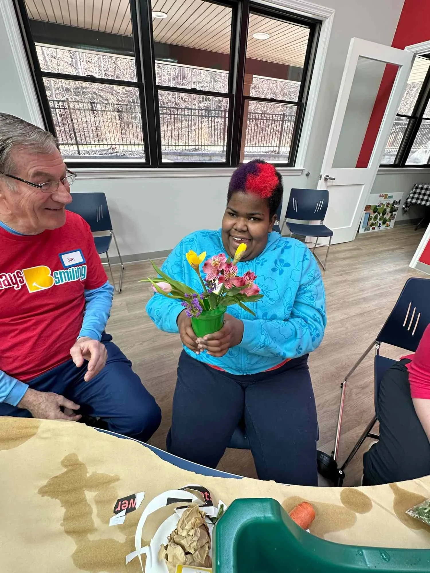 A young woman with short hair dyed with red streaks, smiling and holding a bouquet of colorful flowers, sitting in a room with large windows and a red and white wall. An older man wearing glasses and a red shirt is sitting to her left, smiling at her.