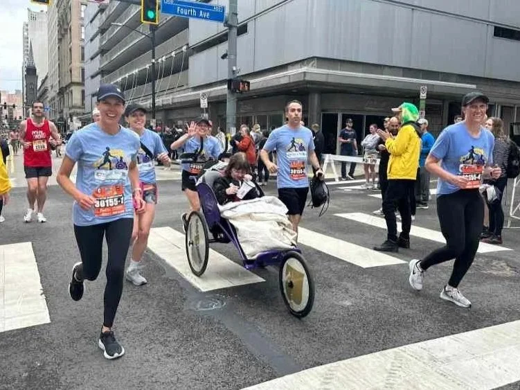 People participating in a marathon, running on a city street with a person in a wheelchair among them. The runners are smiling and wearing blue shirts with race bibs.