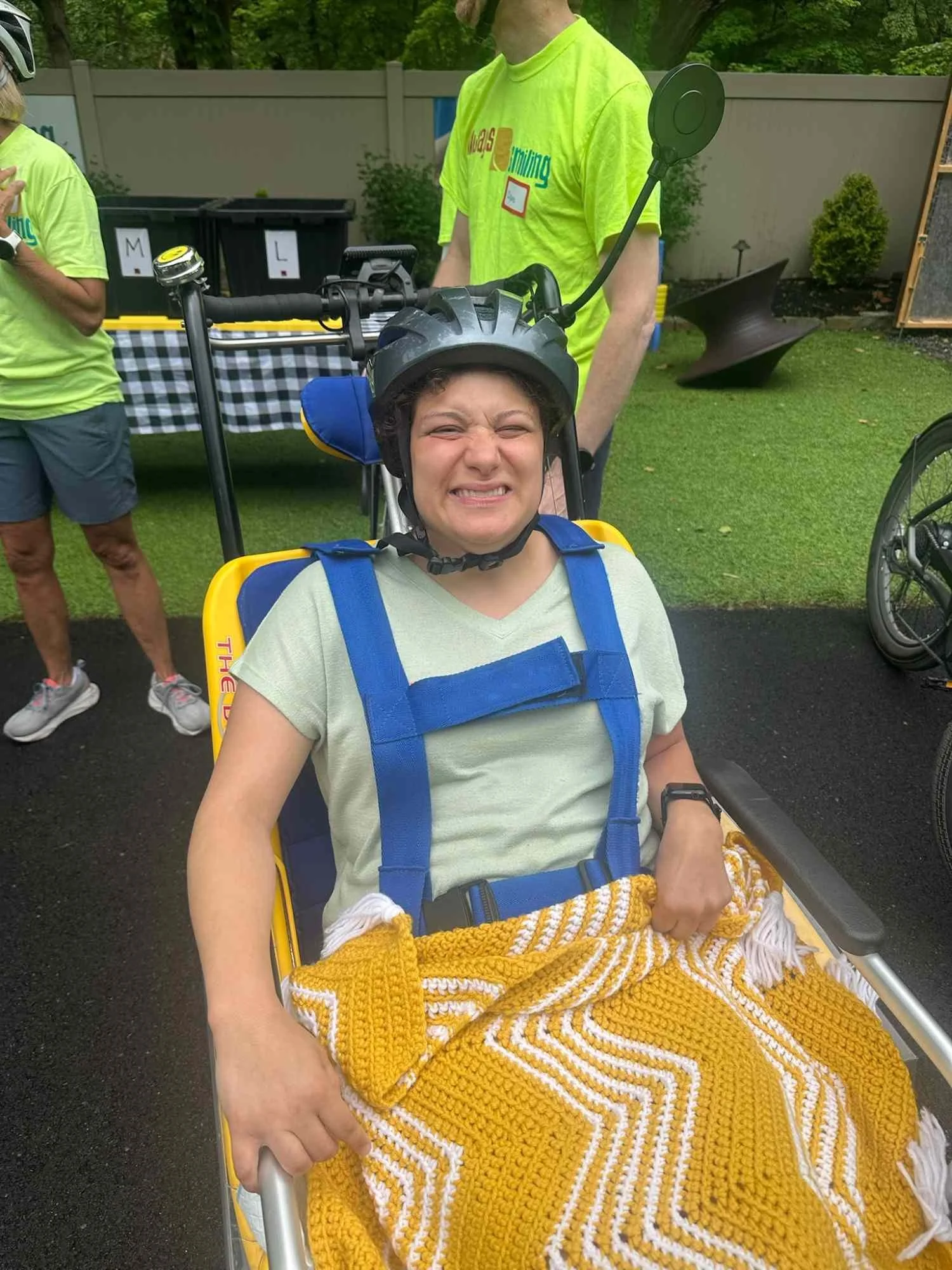 A person wearing a helmet and sitting in a yellow wheelchair, wrapped in a yellow and white knitted blanket, smiling with eyes closed. Two other people are visible in the background, one with a yellow shirt and the other partially visible. The backgr
