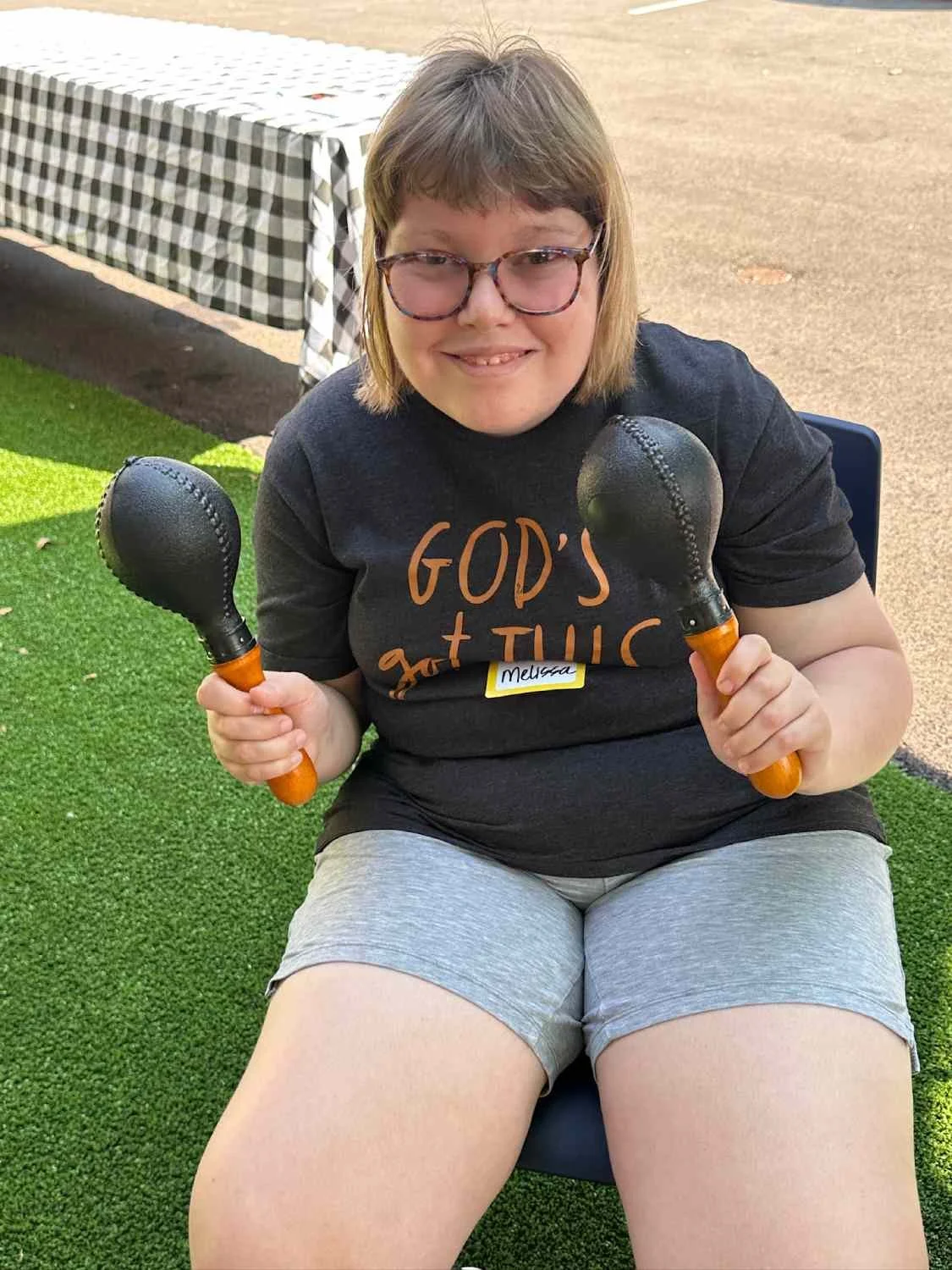 A smiling young woman with Down syndrome, wearing glasses and a black T-shirt that says 'GOD'S GOT THIS' in orange letters, sitting outdoors on artificial grass, holding two black percussion mallets.