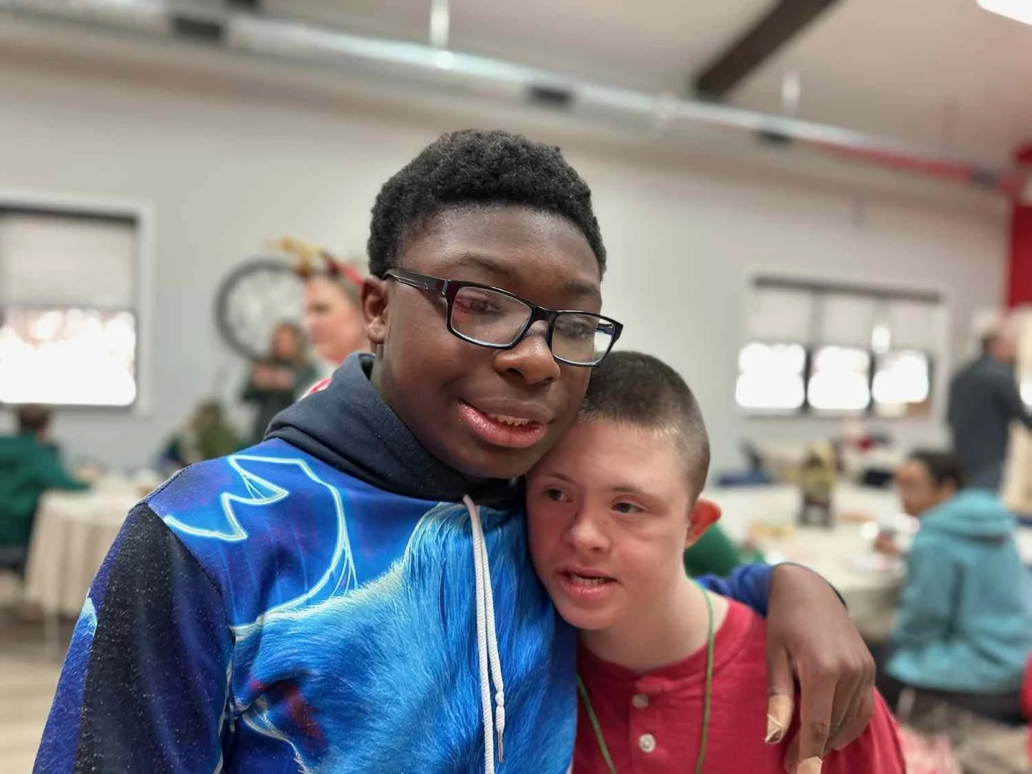 Two young boys embracing, one African American with glasses and a blue hoodie, the other Caucasian with short hair and a red shirt, in a room with other people in the background.