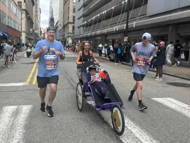 Three runners jogging on a city street during a marathon, with a woman pushing a stroller with an elderly person inside. Buildings and spectators line the street.
