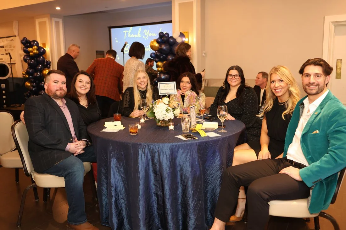 A group of seven people sitting around a decorated dinner table at a celebration event, with balloons and a 'Thank You' message on a screen in the background.