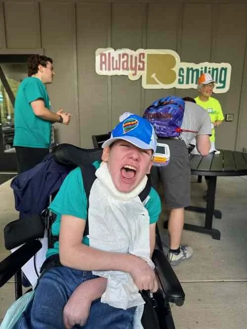 A young boy with a prosthetic leg, wearing a cap and smiling widely, sitting in a wheelchair with a backpack, in front of a sign that says 'Always Smiling', with other children in the background.