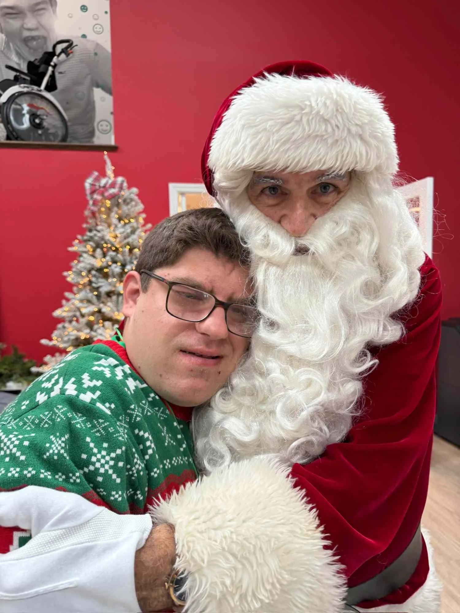 A man in a green Christmas sweater and glasses is hugging Santa Claus, who is dressed in traditional red and white with a bushy white beard. In the background, there is a decorated Christmas tree with lights and a ribbon, and a red wall with a black-