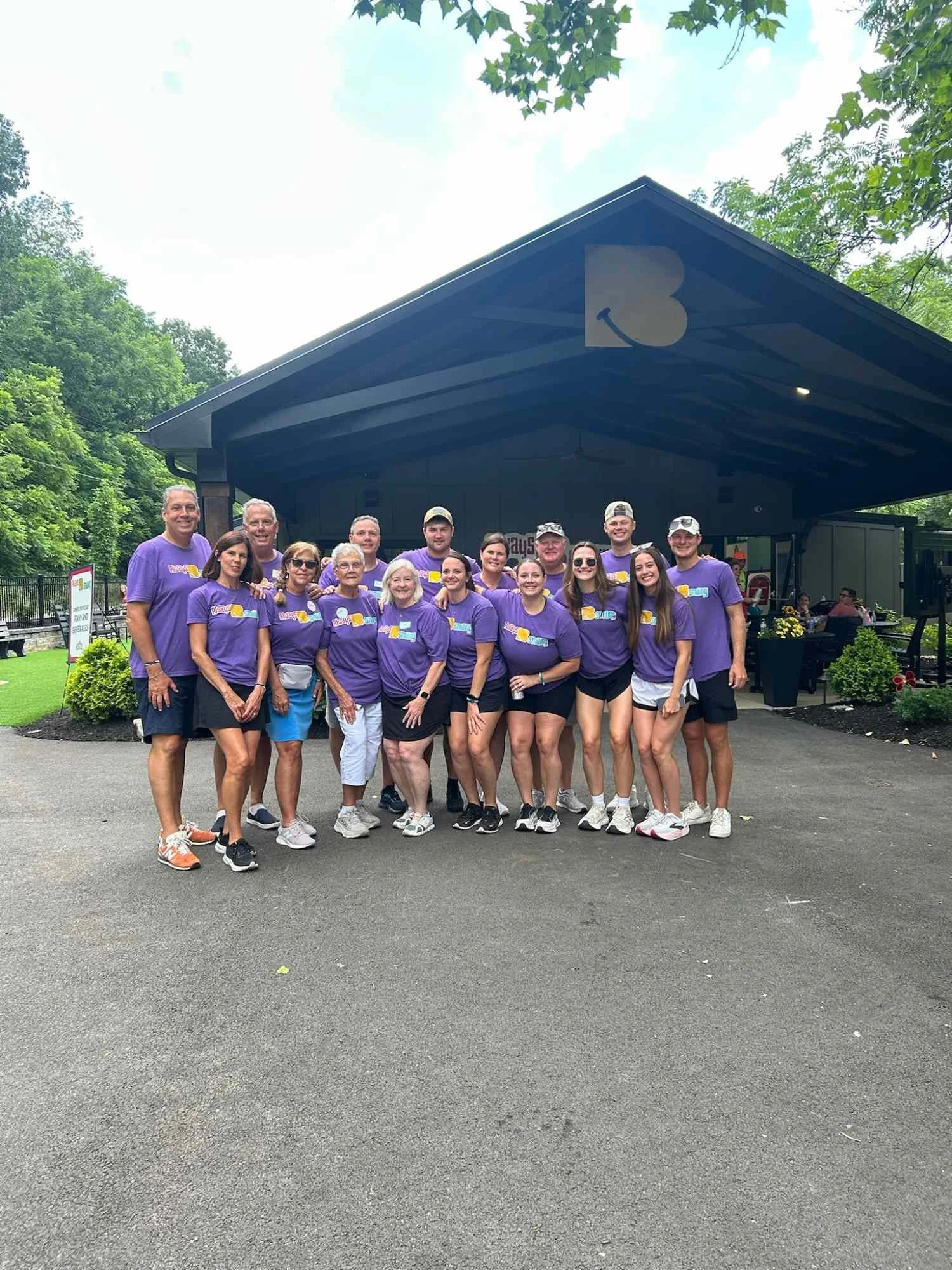 A group of 16 people wearing matching purple T-shirts with a yellow and blue design, standing outside in front of a building with a dark roof and a white sign that says "Days". Some people are wearing sunglasses and hats, and they are posing together