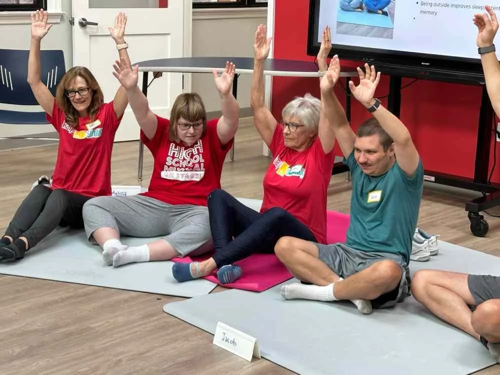 Two women sitting cross-legged on a yoga mat, raising their hands in the air and smiling, in a room with a red and gray wall.
