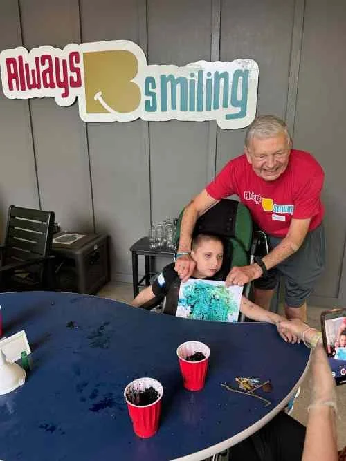 An elderly man with gray hair and a big smile, wearing a red shirt, is standing next to a young boy in a wheelchair. The boy has a serious expression and is holding a colorful picture. There are two red cups with dark liquids on the table, and a pers