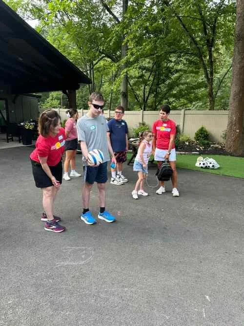Group of children playing volleyball outdoors on a paved area with trees and a fence in the background.