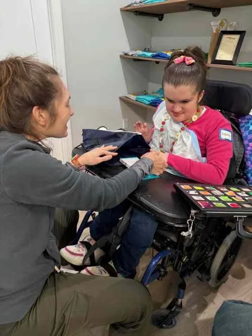 A young girl with a pink bow in her hair is smiling and sitting in a wheelchair. A woman is kneeling in front of her, holding her hand and speaking to her, in a room with shelves and colorful items.