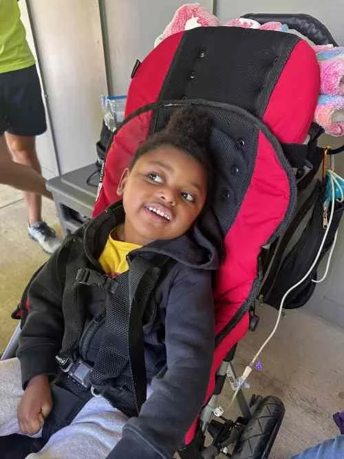 A smiling young boy sitting in a red and black wheelchair with a padded headrest, wearing a black jacket and gray pants, indoors.