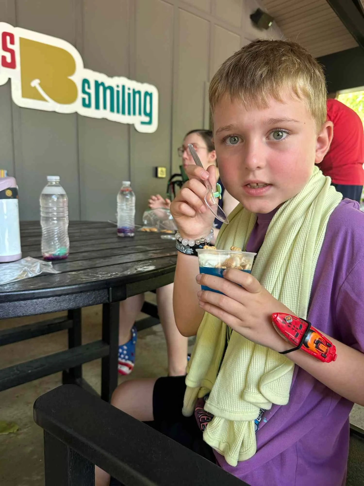 A young boy with green eyes and a red Band-Aid on his wrist sitting at a picnic table inside a covered outdoor area. He is holding a cup of dessert with a spoon and looking at the camera. There are water bottles, a pink container, and a person in the