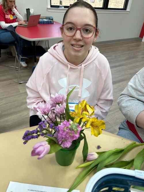 A girl with glasses sitting at a table with a colorful bouquet of flowers in a green pot.