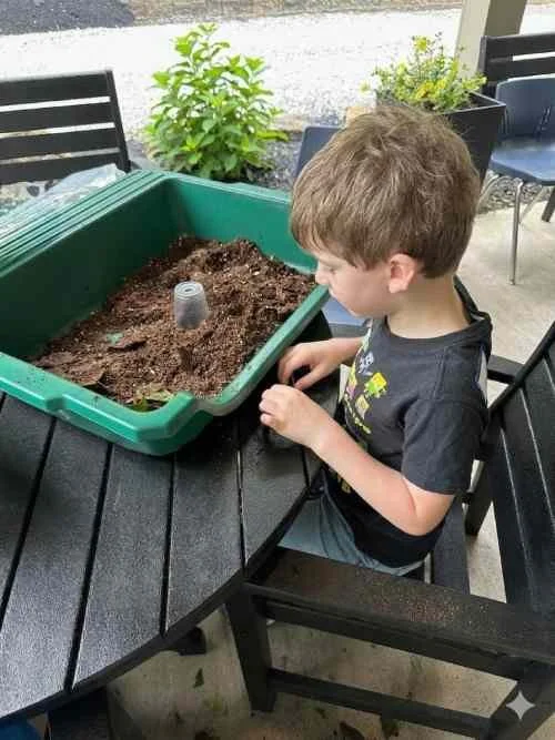A young girl with short brown hair wearing headphones, a white speckled shirt, and blue gloves is sitting at a table. She has a red and white name tag labeled 'Hannah.' In front of her are planting trays with soil and wooden sticks, a red cup, and a water bottle. There is soil spilled on the table, and she is in a room with red, white, and blue furniture, and a window showing an outdoor scene.