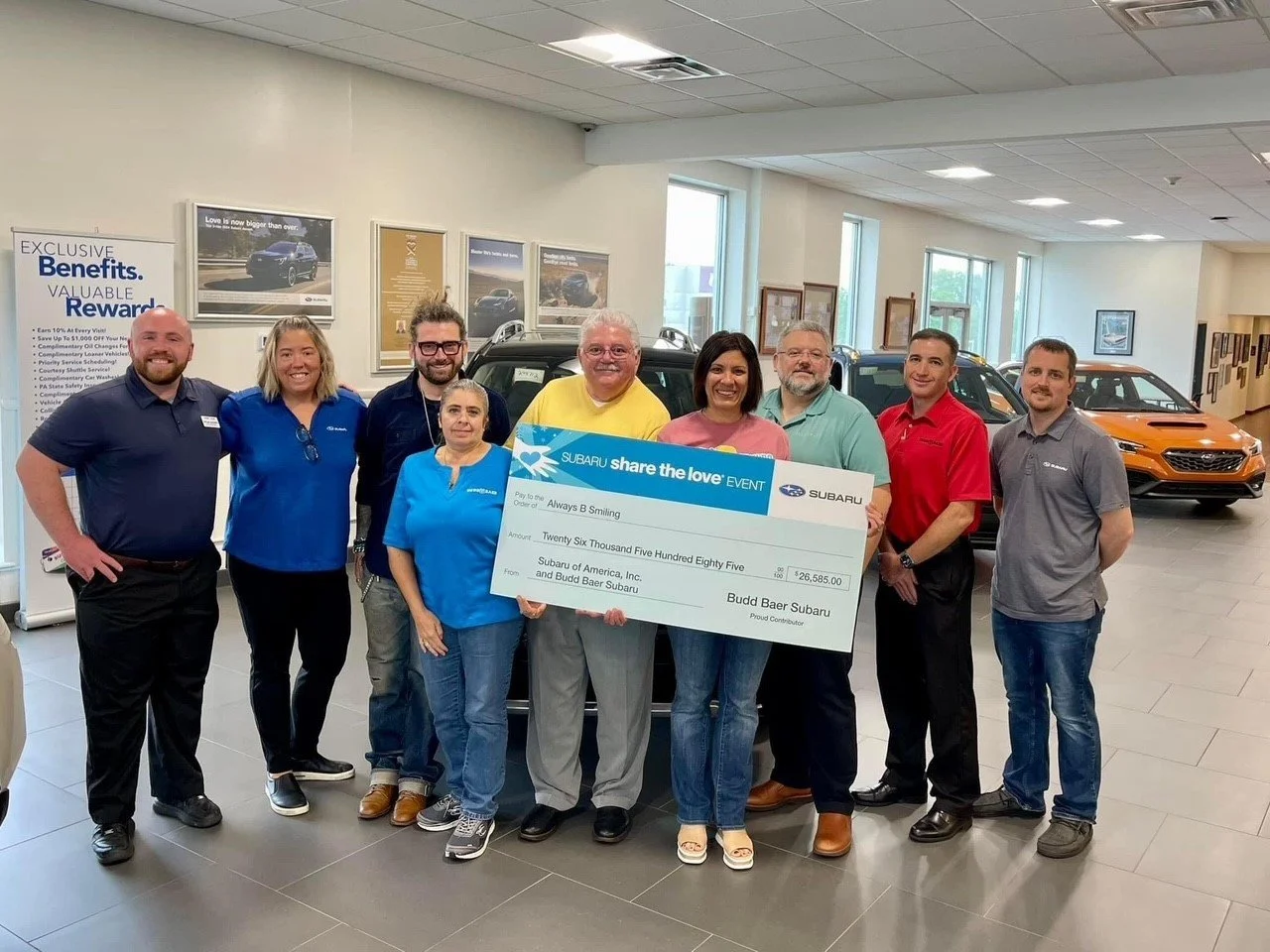 Group of nine people in a car dealership showroom holding a giant check for $26,585, with Subaru cars in the background.