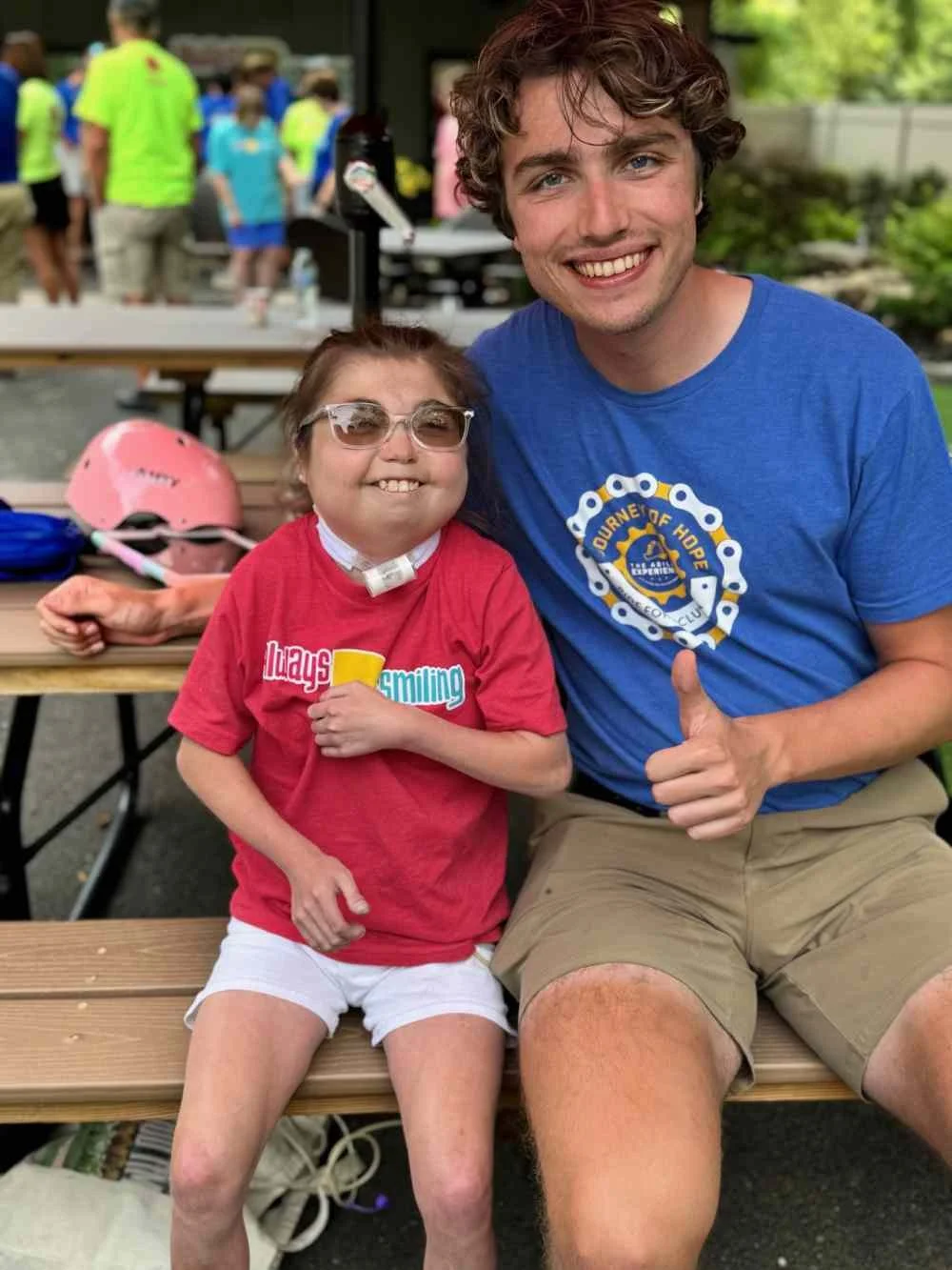 A young girl in a red T-shirt and white shorts, wearing glasses and a neck brace, sitting next to a smiling man in a blue T-shirt with a Rotary Club logo, giving a thumbs-up. They are outdoors at a park or picnic area with other people and tables in the background.