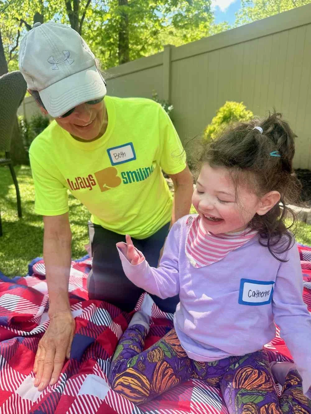 A young girl with curly hair, sitting on a picnic blanket, smiling and pointing, while an older woman in a yellow shirt and white cap, with a name tag 'Beth,' looks on and smiles in a sunny outdoor garden.
