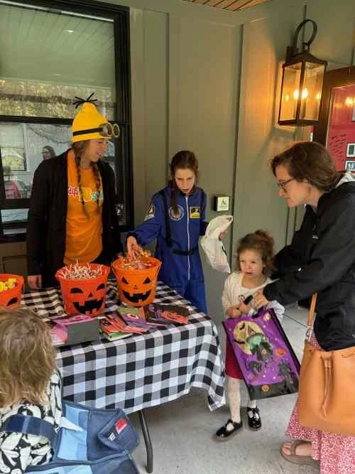 A group of people, including a woman and two young girls, gather at a Halloween-themed table with orange buckets filled with candy. The children are receiving candy from a person dressed in a blue costume, while another person in a black jacket and y