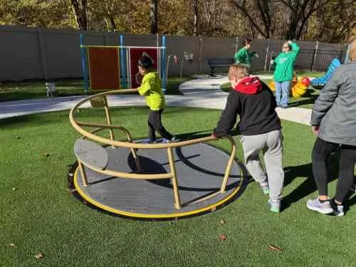 An elderly woman and a young girl sitting on a large, colorful caterpillar-shaped piece of playground equipment, outdoors on grass, with a fence and bee-shaped decoration in the background.