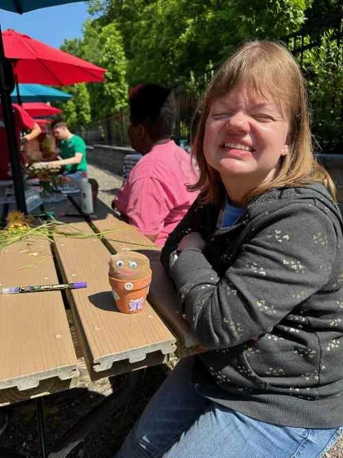 A smiling young girl with brown hair, wearing a gray hoodie, sitting outdoors at a wooden picnic table, with a decorated cupcake with eyes and a smile in front of her. In the background, other children are sitting at the table under colorful umbrellas on a sunny day.