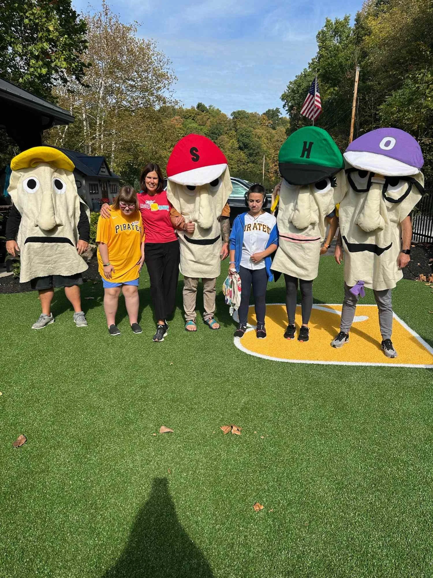 People dress up in mascot costumes resembling ice cream cones with different colored hats and letters on them, standing on a green artificial turf field outdoors. There are five costumed characters and three individuals, one of whom is a woman wearin