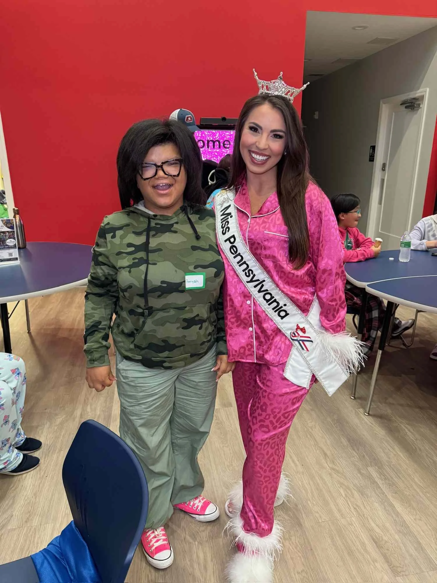 A woman wearing a pink pajama set with animal print and fluffy white slippers, smiling and wearing a crown, standing next to a girl in camouflage hoodie, glasses, and pink sneakers, in a room with a red wall and other children sitting at tables.