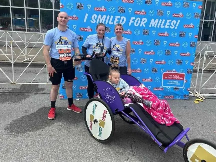 Three adults and a young girl in a stroller at a race event, standing in front of a blue backdrop that says 'I SMILED FOR MILES!'